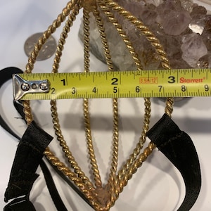 May include: A gold-toned metal headband with a braided design and black elastic straps. The headband measures approximately 2 inches in width.