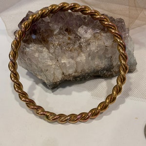 May include: A gold-toned, twisted metal bangle bracelet. The bracelet has a rope-like design and is displayed on a white surface with a crystal formation in the background. The bracelet is circular and appears to be made of metal.