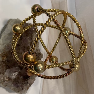 Puede incluir: Un brazalete dorado con un diseño de alambre trenzado y múltiples cuentas redondas. El brazalete está sobre una superficie blanca.