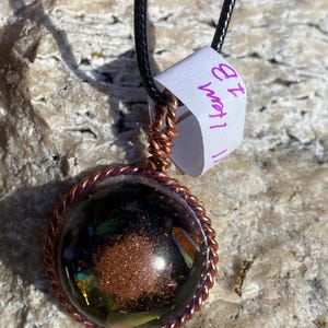 May include: A handcrafted pendant necklace featuring a clear, spherical orb with a dark interior and copper-colored accents. The orb is encased in a copper wire frame and hangs from a black cord. The pendant has a tag with the text "Item 1B".