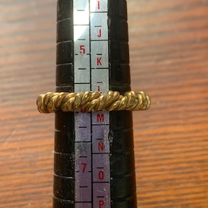 May include: A gold-colored ring with a twisted rope texture positioned on a black ring sizing mandrel. The ring rests between the size 6 and 7 markings on the metal scale, which also displays alphabetical sizing letters from H to Q.