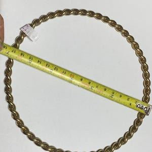 Op de afbeelding: Een goudkleurige, gedraaide metalen ring wordt gemeten met een gele meetlint. De ring heeft een diameter van ongeveer 43 cm. De ring ligt op een witte ondergrond.
