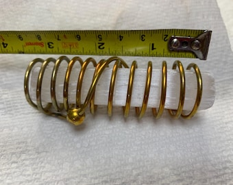 May include: A selenite crystal wrapped in a gold-colored metal coil. The crystal is approximately 3 inches long. The metal coil has a decorative ball at one end. The item is on a white surface.