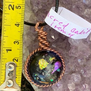 May include: A pendant necklace with a dark, iridescent orb encased in copper wire. The orb has colorful flecks and hangs from a black cord. The tag reads "sacred cabit item". The pendant is approximately 2.5 cm in diameter.