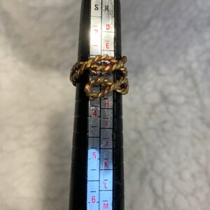 May include: A gold-toned ring with a braided design, measured on a ring sizer that indicates a size 6.