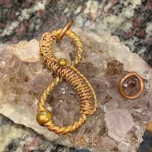 May include: A gold-colored wire-wrapped pendant with two loops and two beads, resting on a bed of amethyst crystals. A smaller copper ring is also visible in the image.