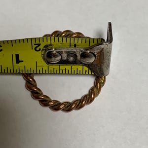 Puede incluir: Primer plano de un anillo de alambre trenzado de color cobre. El anillo tiene un diámetro de aproximadamente 2,5 cm y se mide con una cinta métrica amarilla.