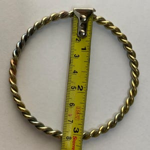 May include: A gold-toned metal bangle with a twisted rope texture. A yellow measuring tape is positioned across the center of the bangle, showing an interior diameter of approximately 2.5 inches from the inner edge to the opposite inner edge.
