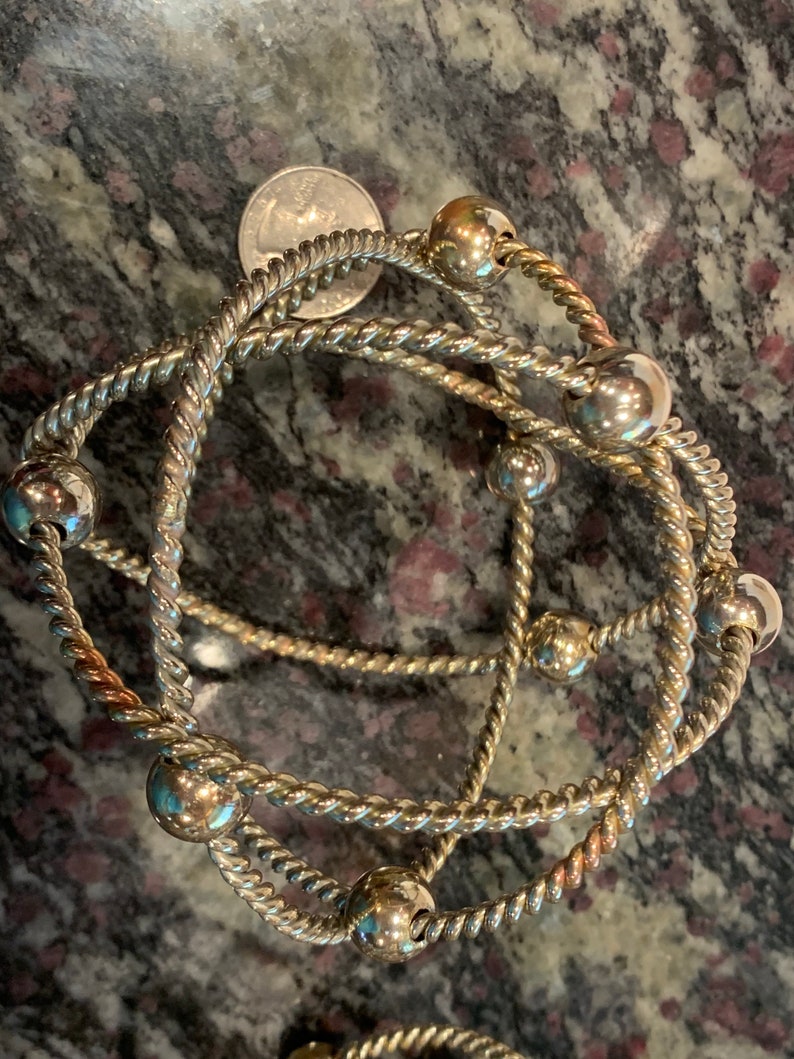 May include: Three silver bracelets with twisted rope designs and silver beads. The bracelets are layered on top of each other.