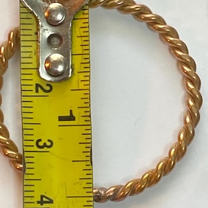 Może przedstawiać: Złoty, skręcony metalowy bransoletka. Bransoletka jest mierzona żółtą miarką, pokazując średnicę około 3,8 cm. Bransoletka ma wzór przypominający linę.