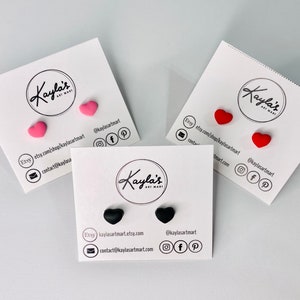 May include: Three pairs of heart-shaped stud earrings displayed on white cards. The earrings are pink, red, and black.
