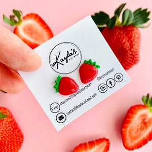 Peut inclure: Une paire de boucles d'oreilles en forme de fraises rouges et vertes sur une carte blanche avec le texte "Kaylas Art Mart" et des coordonnées. La carte est entourée de fraises coupées.