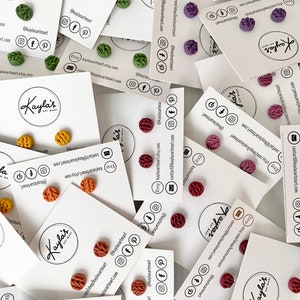 May include: An assortment of handmade polymer clay earrings in various colours, including green, orange, red, and purple. Each pair features a small, textured, circular design. The earrings are displayed on white cards with the Etsy shop name "Kayla's Art Mart" and contact information.