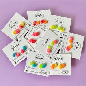 Peut inclure: Assortiment de boucles d'oreilles en forme d'oursons en gélatine de différentes couleurs, présentées sur des cartes blanches avec le texte "Kayla's Art Mart" et des coordonnées. Les boucles d'oreilles sont de couleurs variées, notamment rose, jaune, orange et vert.