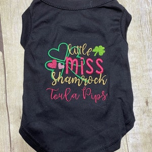 May include: Black sleeveless dog shirt with green shamrock and pink hearts embroidery. The text "little miss shamrock Toula Pips" is embroidered on the shirt.