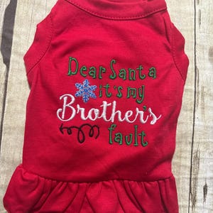 May include: A red pet shirt with a ruffled hem featuring embroidered text that reads Dear Santa it's my Brothers fault in green and white thread, accented by a blue glittery snowflake icon and a black decorative swirl design.