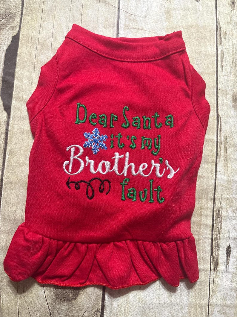 May include: A red pet shirt with a ruffled hem featuring embroidered text that reads Dear Santa it's my Brothers fault in green and white thread, accented by a blue glittery snowflake icon and a black decorative swirl design.