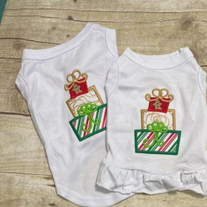 May include: Two white dog shirts with a Christmas-themed design of stacked presents with red, green, and gold embroidery.