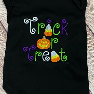 K&ouml;nnte beinhalten: Schwarzes Hunde-T-Shirt mit den gestickten Worten "Trick or Treat" in Wei&szlig;, Gr&uuml;n und Lila. Das Shirt zeigt K&uuml;rbis- und Candy-Corn-Designs, ideal f&uuml;r Halloween.