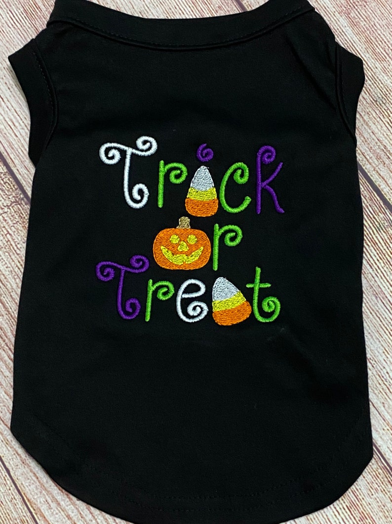 K&ouml;nnte beinhalten: Schwarzes Hunde-T-Shirt mit den gestickten Worten "Trick or Treat" in Wei&szlig;, Gr&uuml;n und Lila. Das Shirt zeigt K&uuml;rbis- und Candy-Corn-Designs, ideal f&uuml;r Halloween.