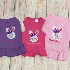 May include: Three sleeveless dog dresses with embroidered bunny faces and names. The dresses are purple, pink, and red. The bunnies have purple bows.