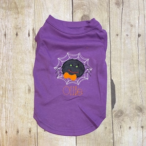 Puede incluir: Camiseta morada para perro con una araña negra bordada que lleva una pajarita y el nombre "Ollie" en hilo naranja.