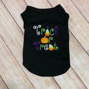 K&ouml;nnte beinhalten: Schwarzes Haustier-T-Shirt mit den gestickten Worten "Trick or Treat" in Wei&szlig;, Gr&uuml;n und Lila, zusammen mit einem K&uuml;rbis und Zuckermais. Das &auml;rmellose Shirt hat einen abgerundeten unteren Saum.