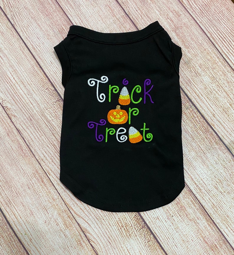 K&ouml;nnte beinhalten: Schwarzes Haustier-T-Shirt mit den gestickten Worten "Trick or Treat" in Wei&szlig;, Gr&uuml;n und Lila, zusammen mit einem K&uuml;rbis und Zuckermais. Das &auml;rmellose Shirt hat einen abgerundeten unteren Saum.