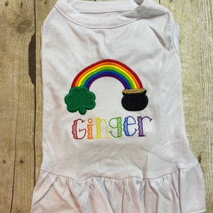 May include: White dog shirt with a rainbow, pot of gold, and shamrock embroidery. The shirt also has the name "Ginger" embroidered on it.