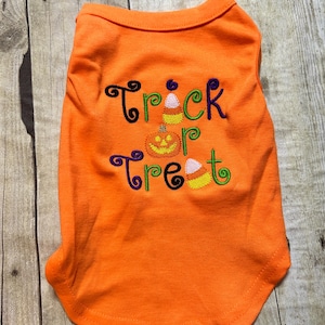 K&ouml;nnte beinhalten: Oranges Hundeshirt mit den gestickten Worten "Trick or Treat" in Schwarz, Gr&uuml;n, Lila und Gelb. Das Design umfasst einen K&uuml;rbis und Zuckermais. Das Shirt ist aus einem weichen Material.