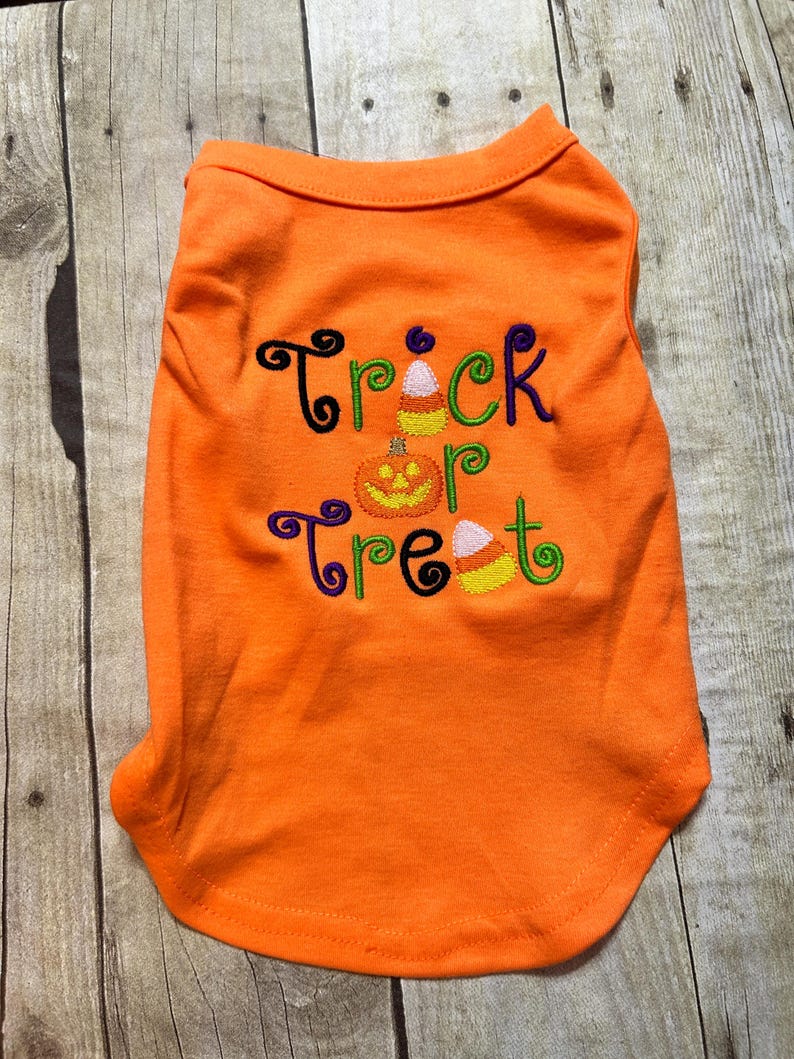 K&ouml;nnte beinhalten: Oranges Hundeshirt mit den gestickten Worten "Trick or Treat" in Schwarz, Gr&uuml;n, Lila und Gelb. Das Design umfasst einen K&uuml;rbis und Zuckermais. Das Shirt ist aus einem weichen Material.