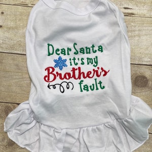 May include: A white pet dress featuring embroidered text that reads Dear Santa it's my Brother's fault in green, red, and black thread. A blue snowflake icon is positioned between the words Dear Santa and it's my. The dress has a ruffled hem.
