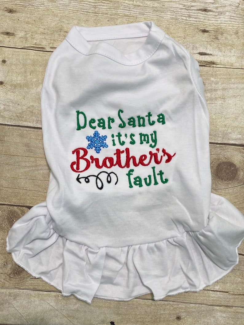 May include: A white pet dress featuring embroidered text that reads Dear Santa it's my Brother's fault in green, red, and black thread. A blue snowflake icon is positioned between the words Dear Santa and it's my. The dress has a ruffled hem.
