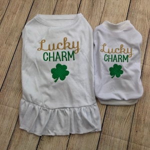 May include: Two white dog shirts with a gold and green embroidered design that says "Lucky Charm" and a green shamrock. The larger shirt has a ruffle at the bottom.