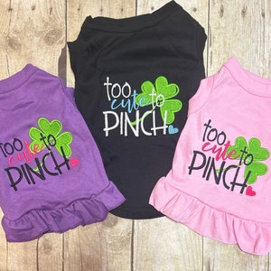 May include: Three dog shirts with a ruffle trim. The shirts are purple, black, and pink. Each shirt has a green shamrock and the text "too cute to pinch".