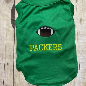 May include: Green sleeveless dog shirt with a brown football and yellow embroidered text that says "PACKERS".