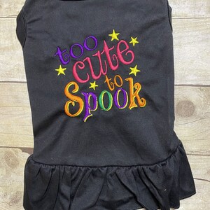 May include: Black dog shirt with a ruffle hem. The shirt has colorful embroidery that says "too cute to spook" with yellow stars.