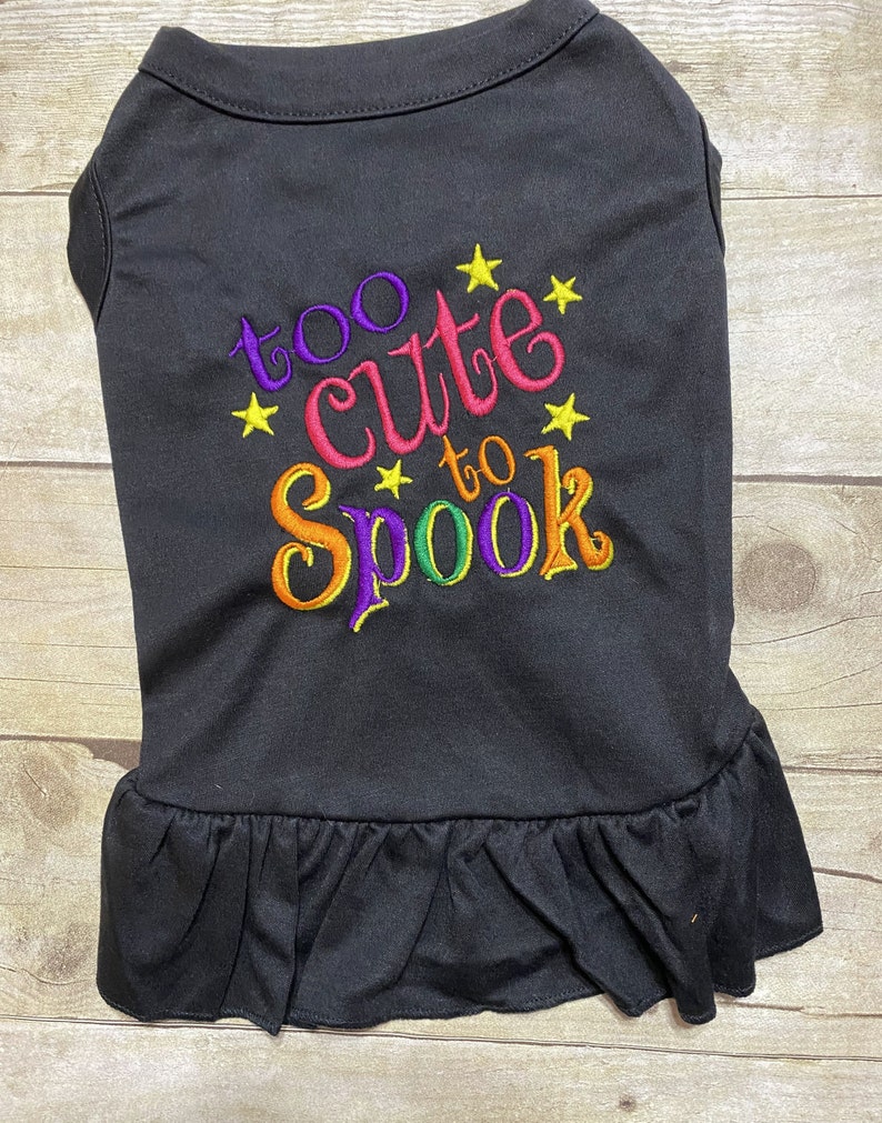 May include: Black dog shirt with a ruffle hem. The shirt has colorful embroidery that says "too cute to spook" with yellow stars.