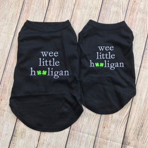 May include: Two black dog shirts with white text that says "wee little hooligan" with three green shamrocks. The shirts are on a wood background.