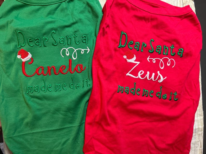 Puede incluir: Dos camisetas para perros rojas y verdes con el texto "Querido Santa [nombre del perro] me hizo hacerlo" y un gr&aacute;fico de gorro de Pap&aacute; Noel. Las camisetas son para Canelo y Zeus.