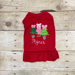 May include: Red dog dress with a ruffle hem and embroidered Christmas trees with pink bows. The dress is personalized with the name "Agnes".