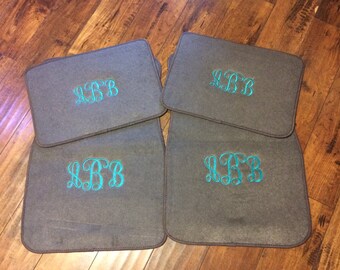 May include: Four gray car floor mats with a teal embroidered monogram of the letters "LBB". The mats are rectangular with rounded corners and a darker brown trim. The mats are arranged on a wooden floor.