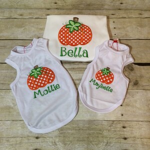 Peut inclure: Trois chemises blanches sans manches avec des citrouilles orange et blanches &agrave; pois et des noms personnalis&eacute;s. Les chemises disent "Bella", "Mollie" et "Maybelle".
