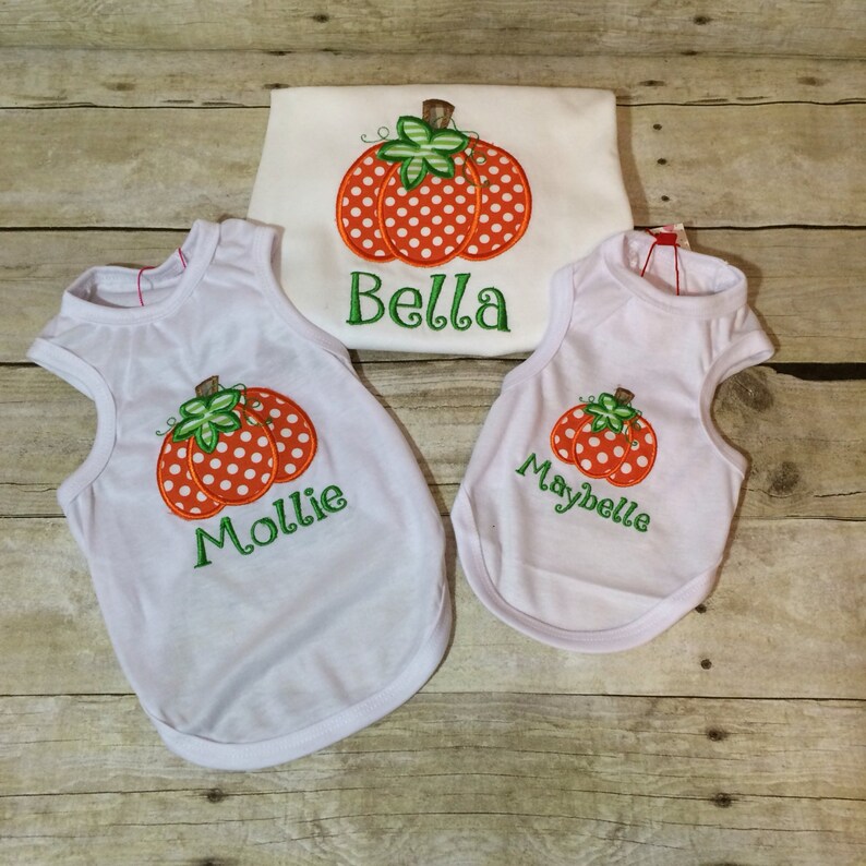 Peut inclure: Trois chemises blanches sans manches avec des citrouilles orange et blanches &agrave; pois et des noms personnalis&eacute;s. Les chemises disent "Bella", "Mollie" et "Maybelle".