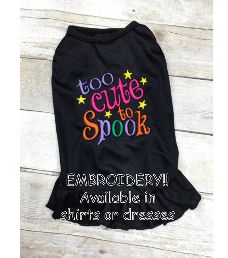 May include: Black dog shirt with colorful embroidery that says "too cute to spook" with yellow stars. The shirt has a ruffled bottom and is available in shirts or dresses.