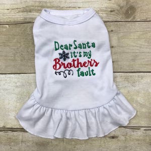 May include: A white pet dress featuring an embroidered design with a black snowflake and the text Dear Santa it's my Brother's fault in green and red thread. The garment has a round neckline and a ruffled hemline at the bottom edge.