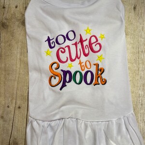 May include: White dog shirt with a ruffled hem. The shirt has colorful embroidered text that says "too cute to spook" with yellow stars.
