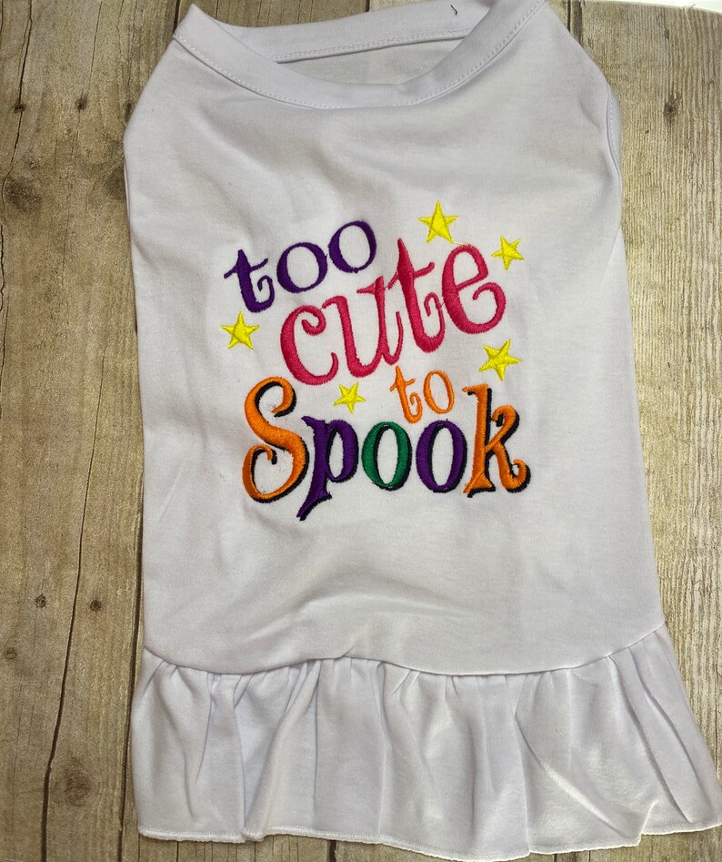 May include: White dog shirt with a ruffled hem. The shirt has colorful embroidered text that says "too cute to spook" with yellow stars.