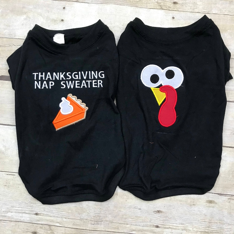 May include: Two black dog sweaters with Thanksgiving themes. One sweater has a pie slice and the text "Thanksgiving Nap Sweater". The other sweater has a turkey face with white eyes and a red wattle.