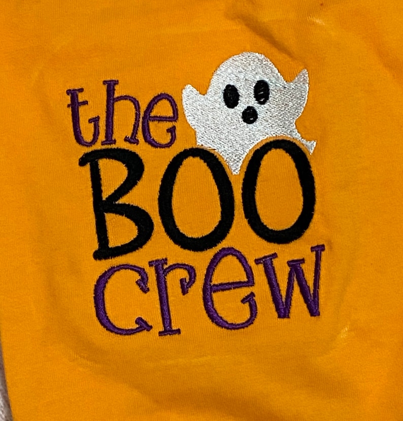 May include: A yellow shirt with the text "the BOO crew" embroidered in black and purple thread. A white ghost with black eyes and a mouth is embroidered above the text.